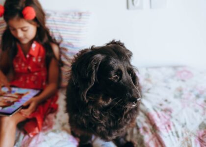 A black dog sits on a bed next to a young girl using a tablet, illustrating the importance of dewormer care in family pets.