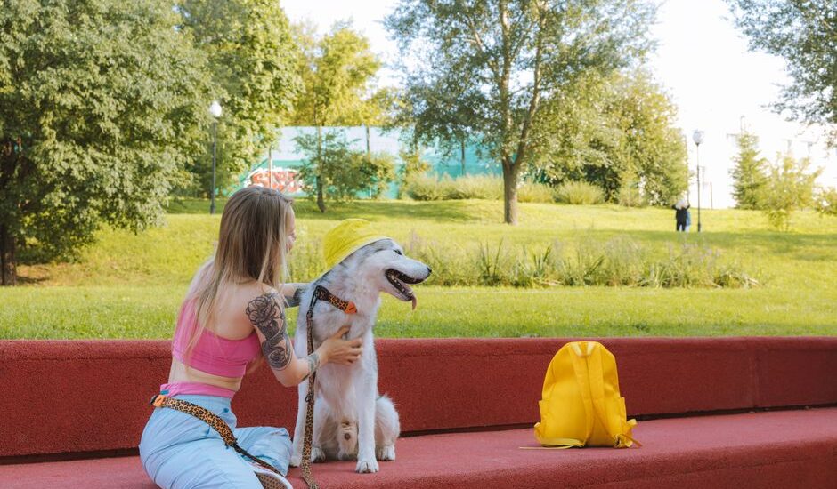 Woman using a hands-free dog leash sits on red steps with her dog, who wears a yellow hat, in a sunny park setting.