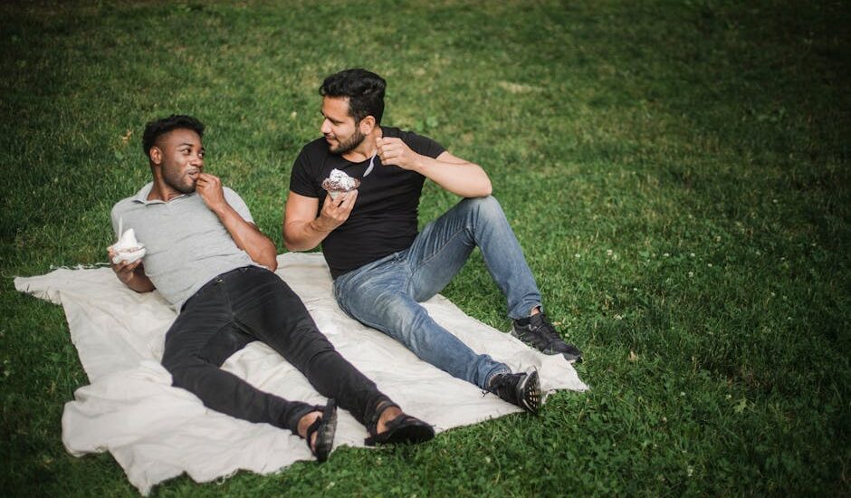 Two men enjoying ice cream on a picnic blanket in a park with a sense of affection and togetherness.