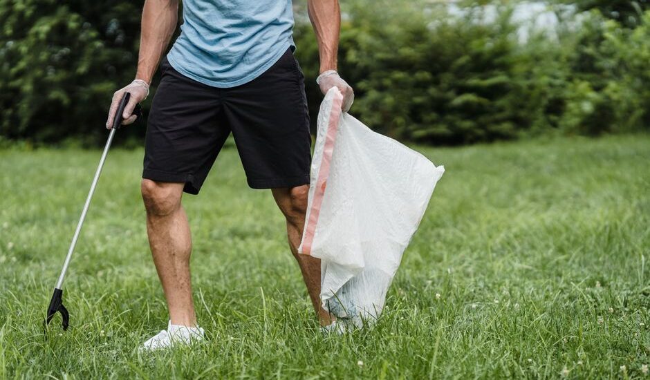 Person cleaning up after a dog walk with a grabber-style poop scooper and plastic bag in a grassy park area.