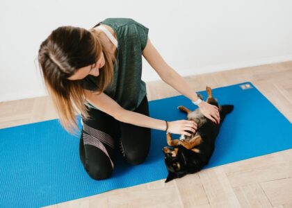 A woman kneels beside a dog on a blue mat, demonstrating how to teach a dog to settle.